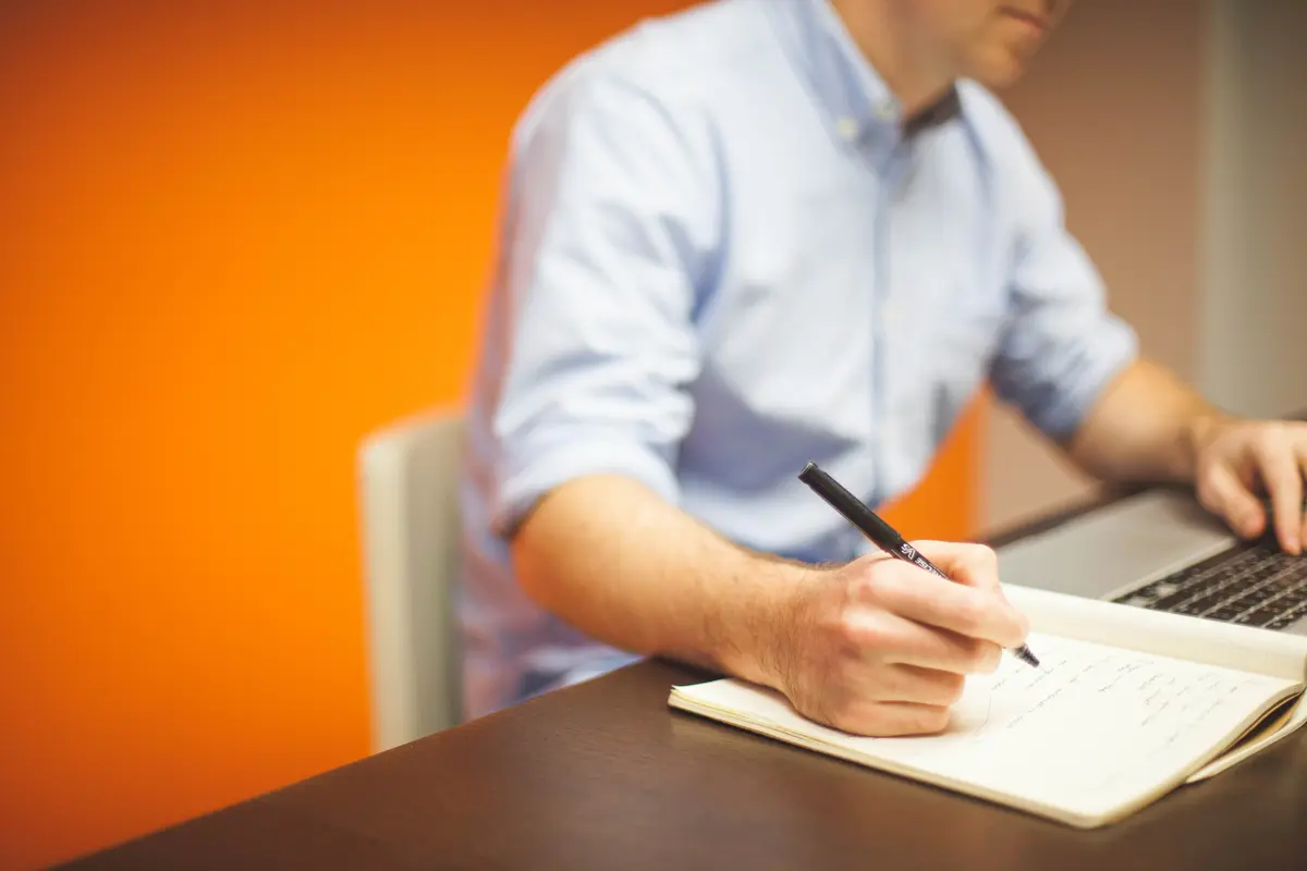 A person writing notes in a notebook beside an open laptop