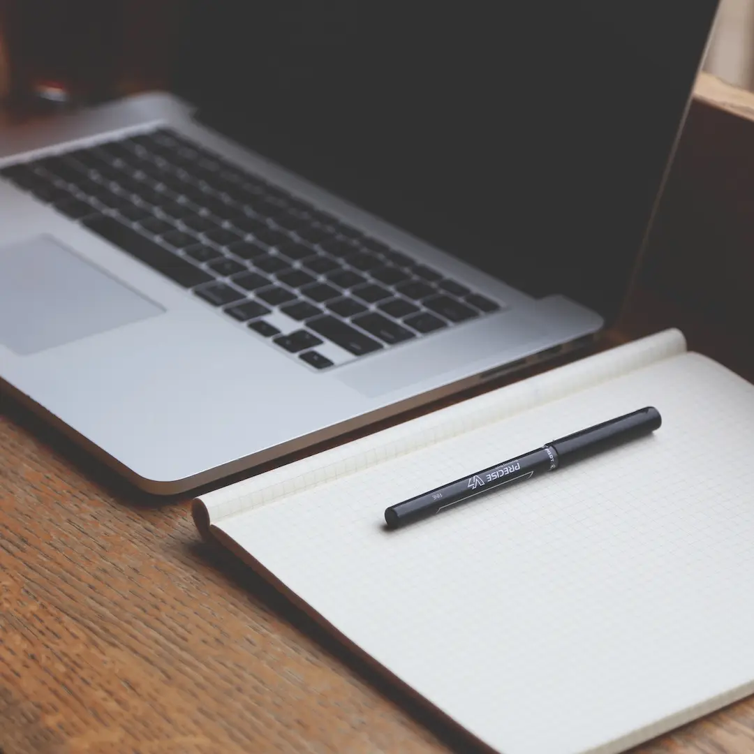 A laptop beside a squared-paper notepad and black pen on a wooden desk